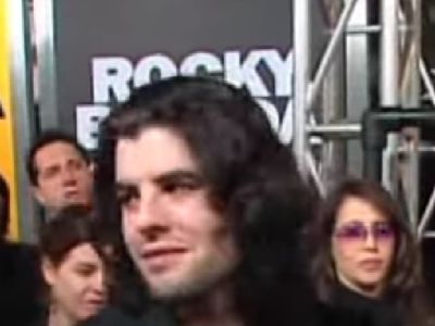Sage Stallone is looking at something as he is surrounded by people.